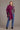 A person wearing a berry color possum merino split front wrap with a mulberry silk detail, paired with a grey sweater, blue jeans, and black boots on a plain gray background