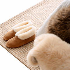 Pair of brown slippers with white sheepskin lining on a textured mat alongside sheepskin rug on a white background
