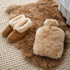Pair of brown slippers with sheepskin lining and a sheepskin water bottle cover, both resting on a sheepskin rug